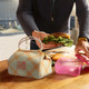 A patterned light green lunch box bag with an attached handle, beside a person holding a sandwich and a pink container.