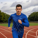 A man in a blue long-sleeved shirt runs on a track, with a logo on his shirt and a cloudy sky above.