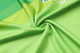 A close-up of a vibrant green fabric, showcasing a smooth texture with a subtle swirl pattern. The material has a logo.