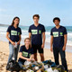 A group of four people wearing navy blue V-collar tee shirts stands on a beach with bags of collected waste.