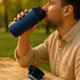 A reusable blue double wall coffee cup with a black lid, set on a table beside a man drinking from it outdoors.