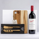 A wooden cheese board with a knife set in a box and a bottle of red wine. The board has a rounded cutout.
