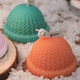 Two hat-shaped candles in green and orange, featuring a textured knit pattern, displayed on a wooden surface with snow.