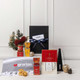 A customised gift set featuring a black box, gourmet treats, a grey towel, and decorative items in red and gold.