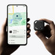 A hand holds a small black Bluetooth tracker next to a smartphone displaying a map and tracking interface.