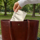 A cream portable makeup bag being placed into a brown leather handbag, set against a green park backdrop.