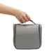 A grey hanging travel toiletry bag with a handle, being held by a hand. It features a zippered closure.