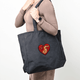 A large black tote bag with a heart-shaped design featuring a dragon, held by a person.