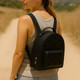 A black EVA Holes Backpack worn on the back of a woman walking along a sandy path in a natural setting.