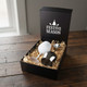 A gift box containing a white egg and a stainless steel wine glass, resting on straw. The box has a festive design.