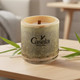 A bamboo candle with a wooden wick, featuring a logo, set on a wooden table with green foliage.