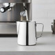 A stainless steel milk jug with a handle, positioned on a countertop near a coffee machine.