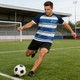 A male model wearing a navy and blue striped as well as white V-neck raglan tee, kicking a soccer ball on a field.