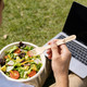 A person holding a wooden fork over a salad bowl, with a laptop in the background on grass. The fork has a logo.