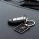 A square silver key ring with a logo, attached to a car key, placed on a black textured surface.
