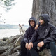 A black windbreaker worn by two people sitting by a tree, with a small dog nearby. The jackets have a logo on them.