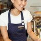 A wooden chalkboard name badge attached to a navy apron worn by a smiling person in a café setting.