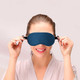 A satin sleep mask in navy blue, being held up by a smiling woman in a light-coloured robe.