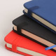 A stack of PU A6 notebooks in navy blue, black, and red, featuring elastic bands and rounded corners.