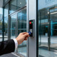 A hand holding a blue NFC keytag approaches a modern glass door with a security reader and indicator lights.