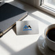 A stainless steel business card with a logo, resting on a table near a notebook and a coffee cup.