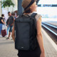 A black convertible travel backpack with a carry handle and shoulder straps, worn on a person's back at a train station.