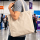 A person holds a beige 450gsm Victorian canvas tote bag with shoulder straps. The bag features a logo.