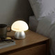 A table lamp with a soft, white dome shade and a silver base, placed on a wooden bedside table with a coffee cup and glasses.