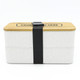 A two-layer lunch box in white with a bamboo lid, secured by a black strap. It features a logo on the top.