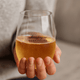 A stemless shatterproof red wine glass filled with sparkling golden beverage, held in a person's hand. Features a logo.
