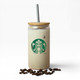 A drink bottle with a bamboo lid and metal straw, featuring a logo and filled with a creamy beverage, placed on coffee beans.
