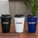 A coffee mug in white with a black lid, featuring a logo, held by a person sitting outdoors.