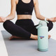 A drink bottle in mint green with a lid and a logo, placed on a yoga mat beside a person in activewear.
