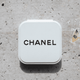 A compact mirror in a white case with a logo on the top, set against a textured stone background.