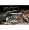 A promotional setup featuring Hornady Precision Hunter 6.5 PRC 143 Gr ELD-X ammunition. The scene includes a rifle equipped with a scope, placed on a textured gun stock, alongside a box of ammunition prominently displayed. The box, matching the cartridges, shows the distinctive red-tipped bullets. Additional hunting gear, like camouflage clothing and a rangefinder, is also visible, setting the context for a hunting or precision shooting scenario.