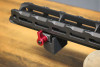 The image showcases the XLR Industries M-LOK Micro Barricade Stop attached to a rifle's handguard, featuring a prominent red quick-detach swivel. This setup demonstrates the practical application of the barricade stop for shooters needing reliable support against various surfaces for steady aim. The red swivel not only provides an aesthetically pleasing contrast to the black handguard but also serves as a functional component for quick sling attachment, facilitating ease of movement and transition for the shooter. The image subtly highlights how such tactical accessories can enhance the functionality and ergonomics of precision rifles.