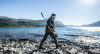 Outdoor scene depicting a hunter traversing a rocky lakeside with a rifle equipped with a GRS Bifrost stock in black, designed for a Bergara B-14 SA HMR. The hunter, in casual outdoor attire, is actively moving across the rugged terrain under bright sunlight, with a majestic lake and mountains in the background. This image captures the essence of an adventurous hunting experience in a stunning natural setting, highlighting the functionality and ergonomic design of the rifle stock.