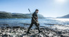 Man walking along a rocky lakeshore carrying a Tikka T1X rifle with a GRS Bifrost stock in green, amidst a scenic backdrop of a lake and mountains under a clear blue sky.