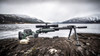 Dramatic outdoor scene featuring a precision rifle equipped with a GRS Bifrost Spigot (104560) mounted on a bipod, set against a rugged lakeside backdrop with snow-covered mountains. The rifle and its accessories, including a GRS stock in green and a black bipod, are displayed on a textured wooden surface, highlighting the equipment’s robustness and readiness for extreme outdoor shooting conditions.