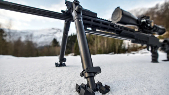 GRS Stocks - GRS Bipod & Adapter Set (105842) in use on a snow-covered ground. Close-up image of a tactical rifle supported by a durable GRS bipod with extended legs and a sturdy adapter set. The bipod is positioned on snowy terrain, demonstrating its stability and grip with clawed feet. The background features a forest and snow-covered mountains, emphasizing the bipod’s utility in harsh, winter conditions and its role in precision shooting.