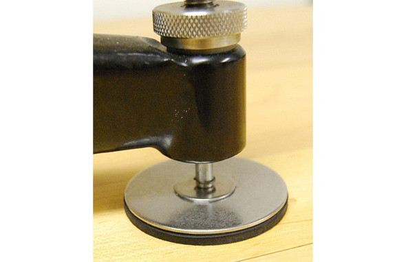 Close-up view of a Benchrite 2-inch Standard Stabilfoot attached to the base of a shooting bench equipment, set on a wooden surface. The metallic, circular stabilizer is designed to enhance steadiness and prevent sliding during use.