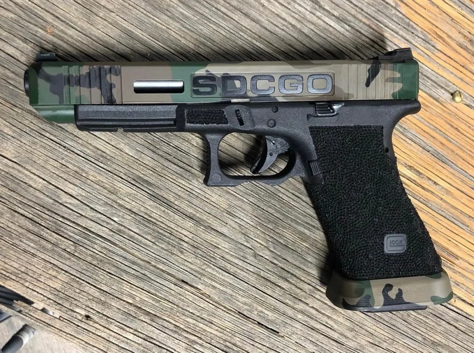 The Glock 34 from the charity auction, shown from one side: a camouflage Cerakote slide with an SDCGO plate, a stippled grip and a camouflage magazine base plate, on a weathered wood table