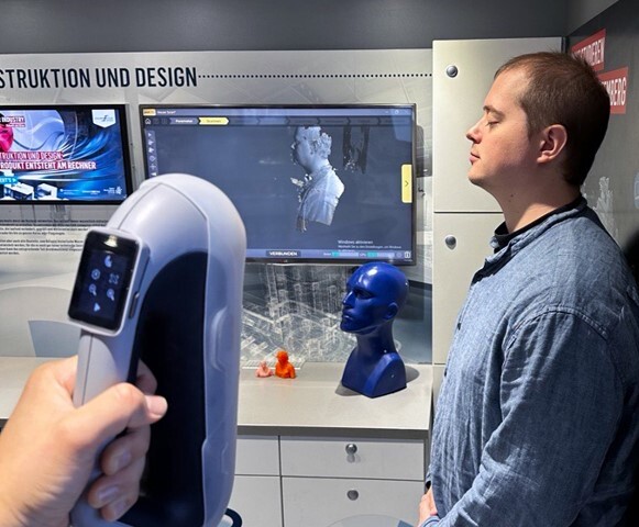 Someone in the Construction and Design station using the Peel 3 scanner to scan the head and torso of a student while the scan is showing on a screen in the background. 