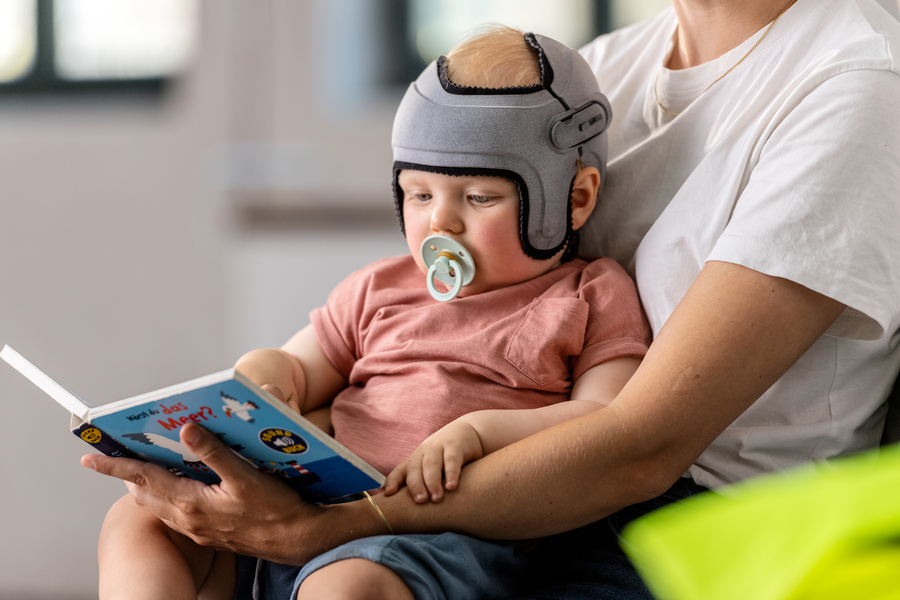 Baby with a pacifier sitting on his mother while reading a book and wearing a grey MyCRO band. 