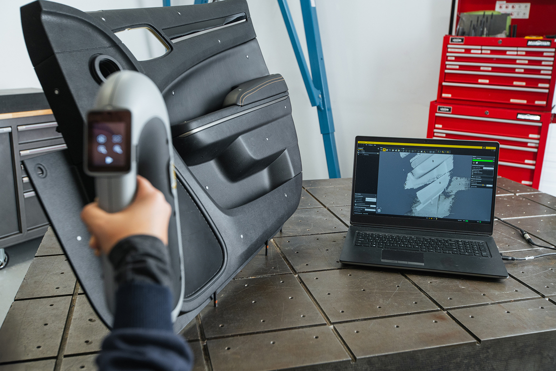 A person using the Peel 3 3D scanner to scan an interior car door panel on a table next to a laptop displaying the scanned data in real time 