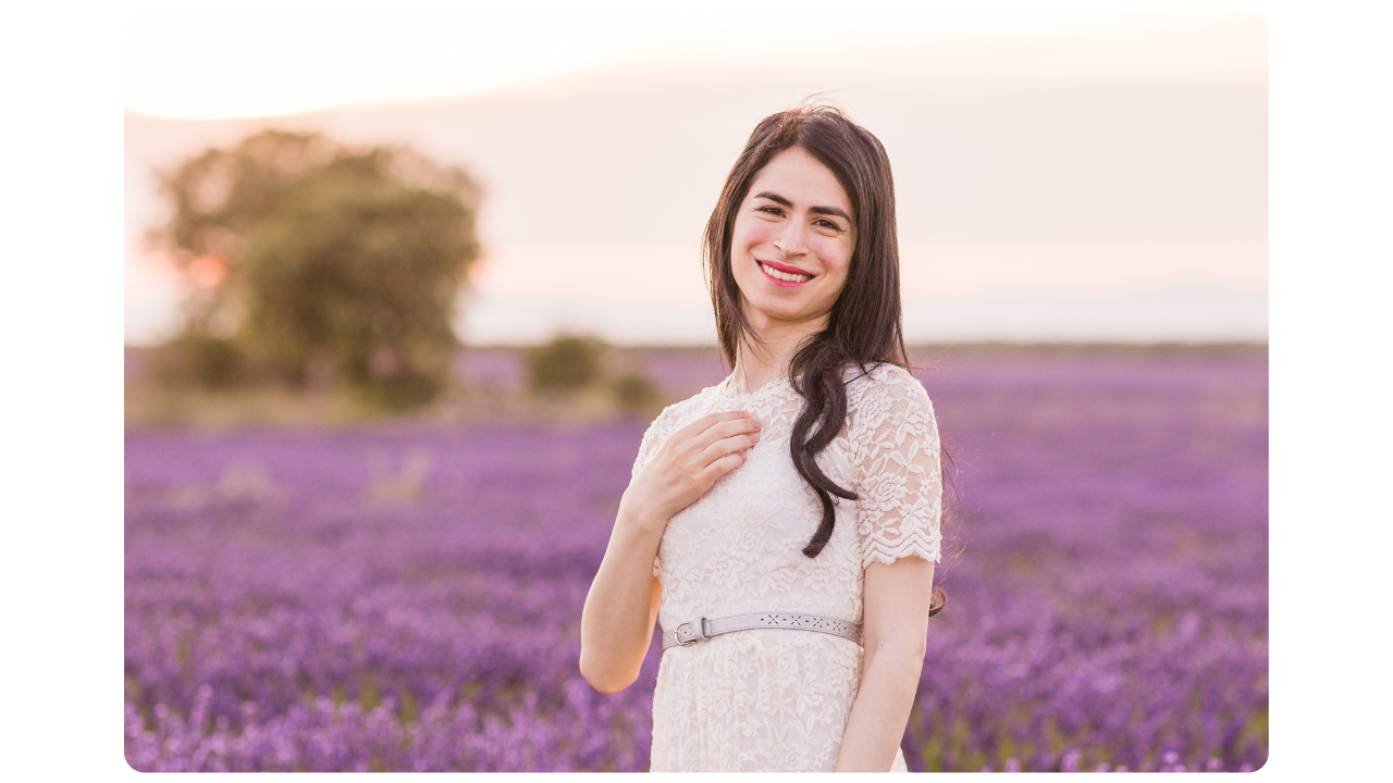 Smiling transgender woman in a white dress standing in a field of purple flowers Gender X Life.com