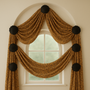 This elegant arched window treatment features golden fabric with black swirl patterns, gracefully swagged on six dark medallions against warm neutral walls for a timeless, sophisticated look.
