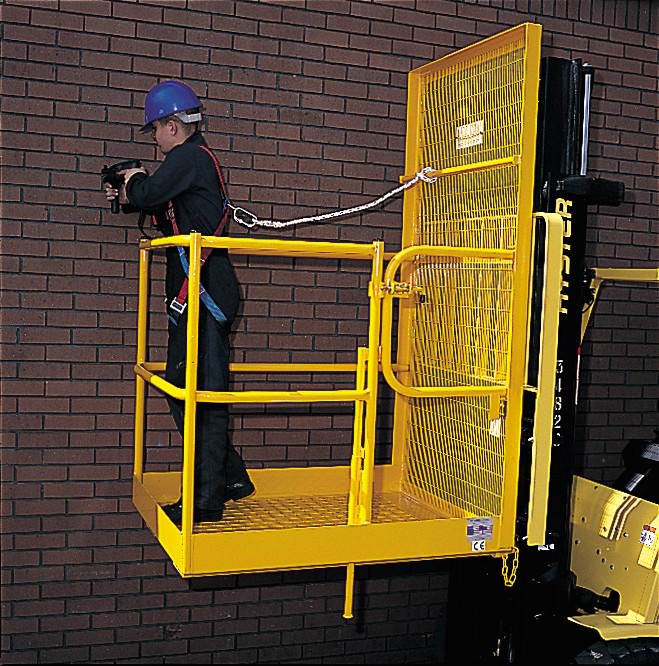 A yellow gated forklift raised access platform with a gate for two people showing someone standing on it, attached to the platform by a harness as they lean over the edge and 