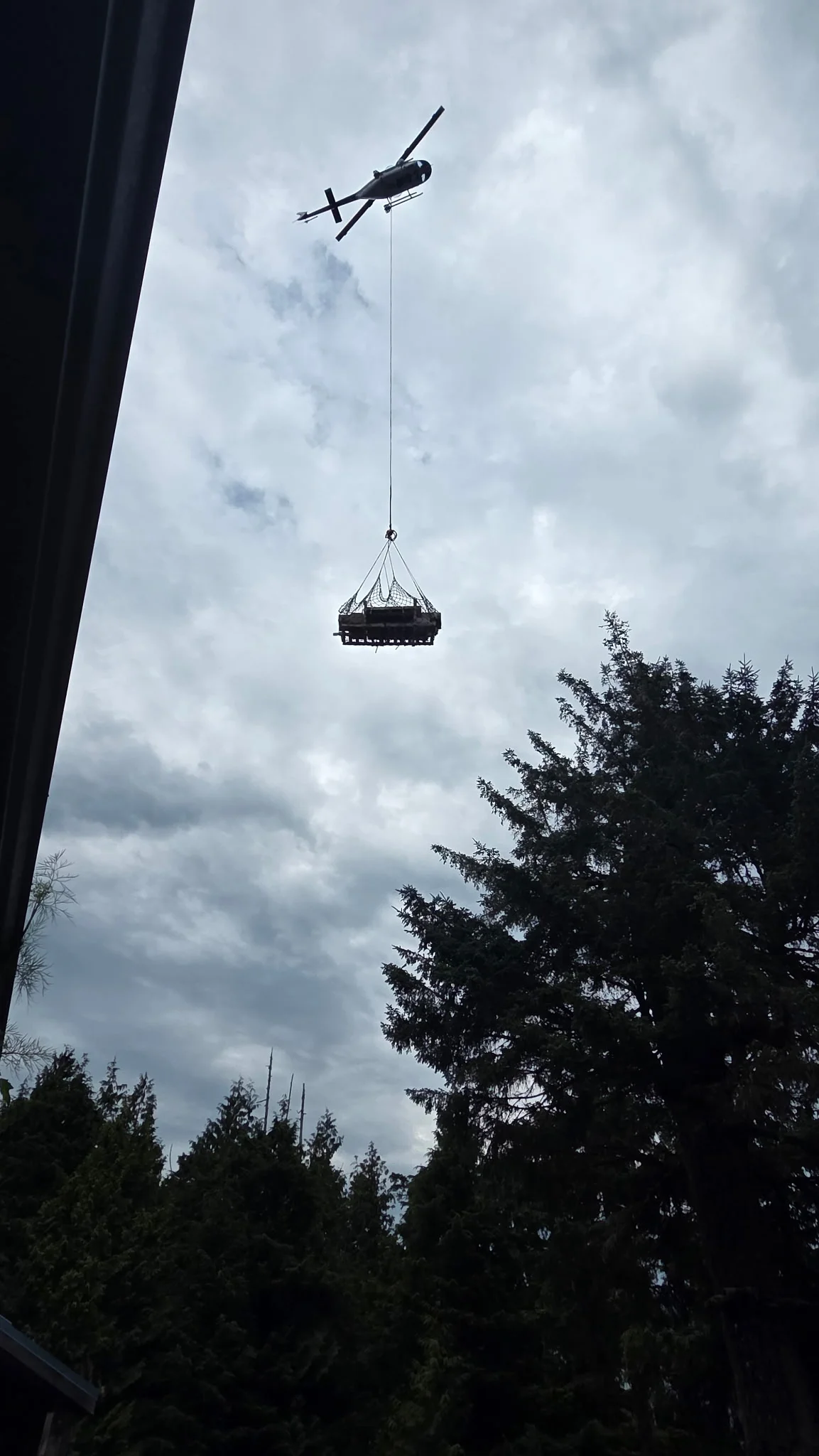 A helicopter carrying a suspended cargo sling above trees during a delivery.