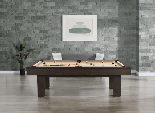 Dark wood pool table with tan felt, billiard balls, and cues in a modern room with gray brick wall.